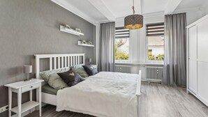 A bedroom with a white bed, grey curtains, a window with blinds, and a radiator. - Cozy apartment in Bad Harzburg with WiFi (Bad Harzburg)