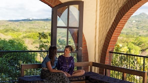 Un balcon où deux personnes sont assises sur un banc, avec vue sur un paysage luxuriant.