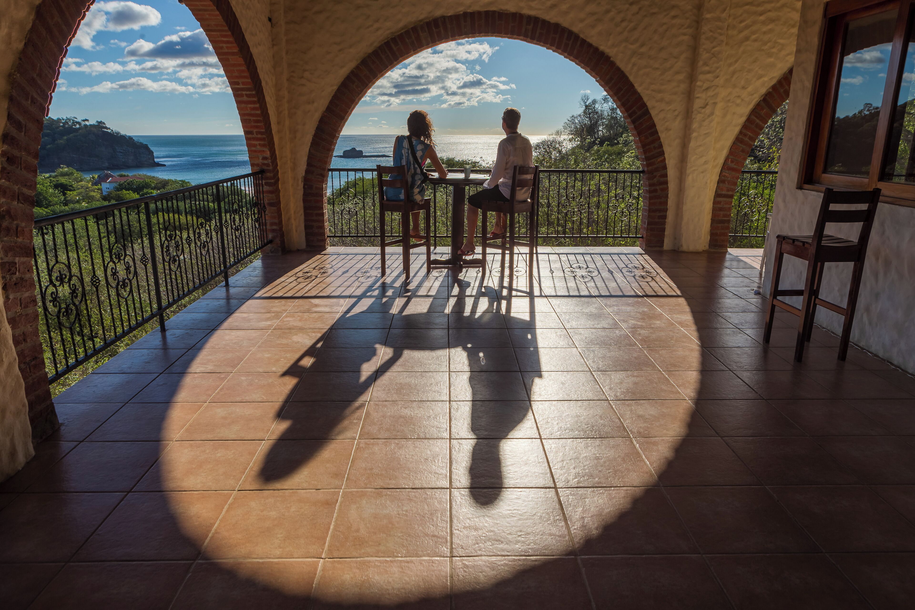 A balcony with two people seated, overlooking a coastal view.