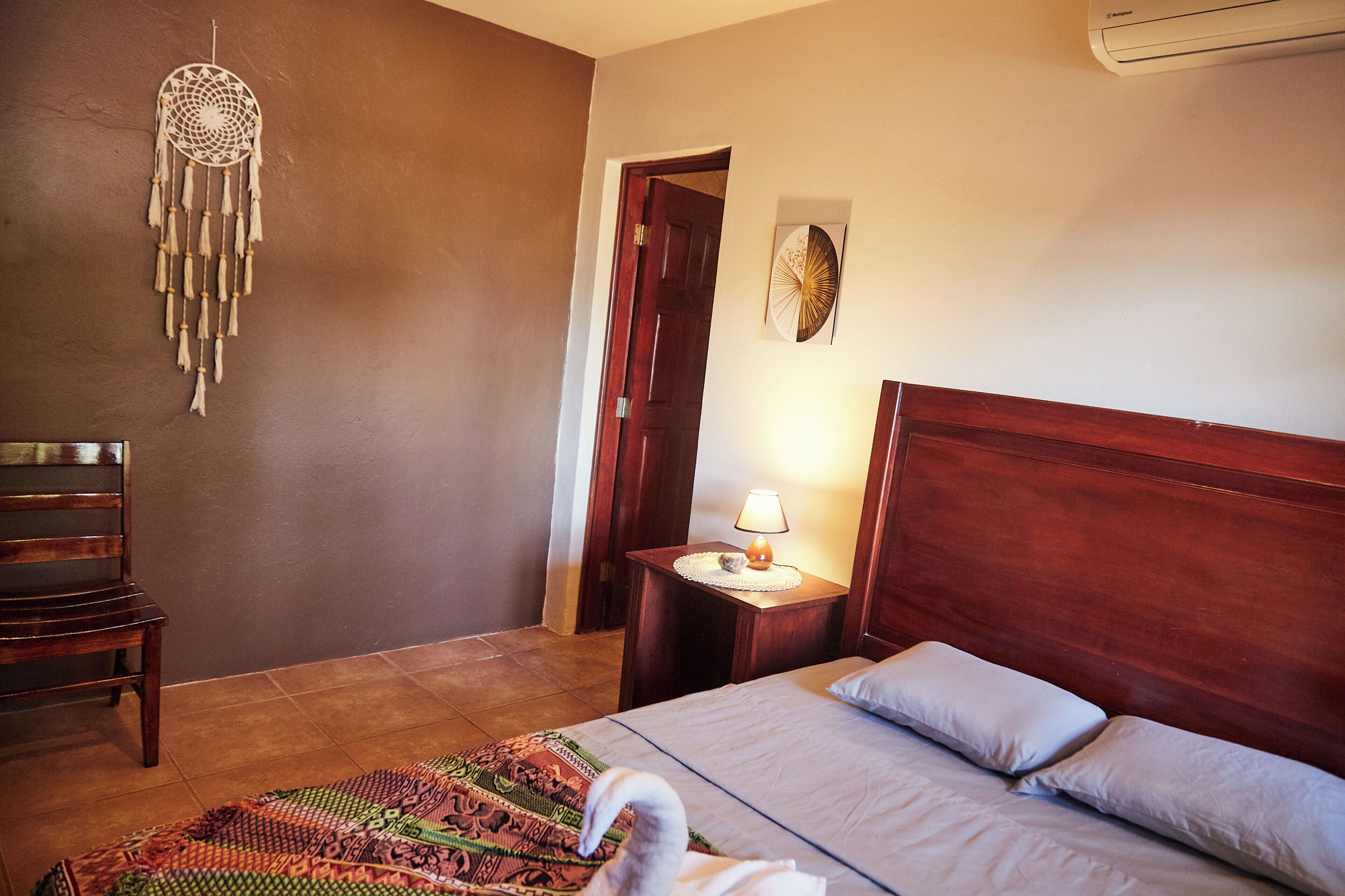A bedroom with a wooden bed, a nightstand with a lamp, a dreamcatcher on the wall, and a chair.