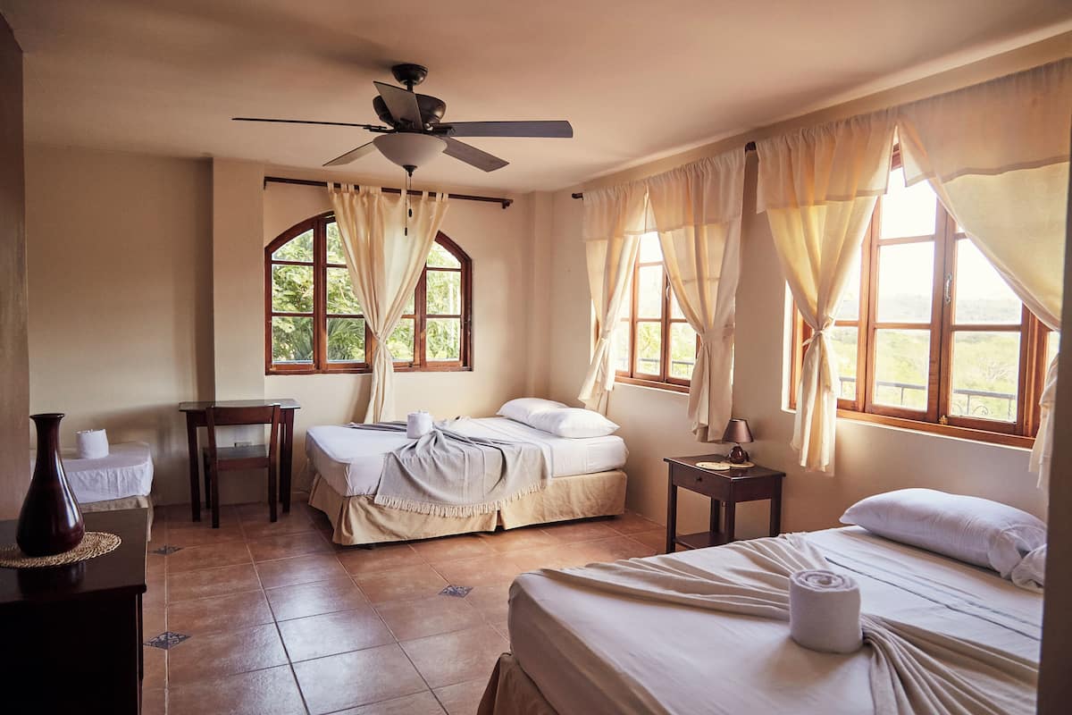 Une chambre d’hôtel avec deux lits, un ventilateur de plafond, une fenêtre avec des rideaux, une petite table et un vase placé sur le côté gauche.