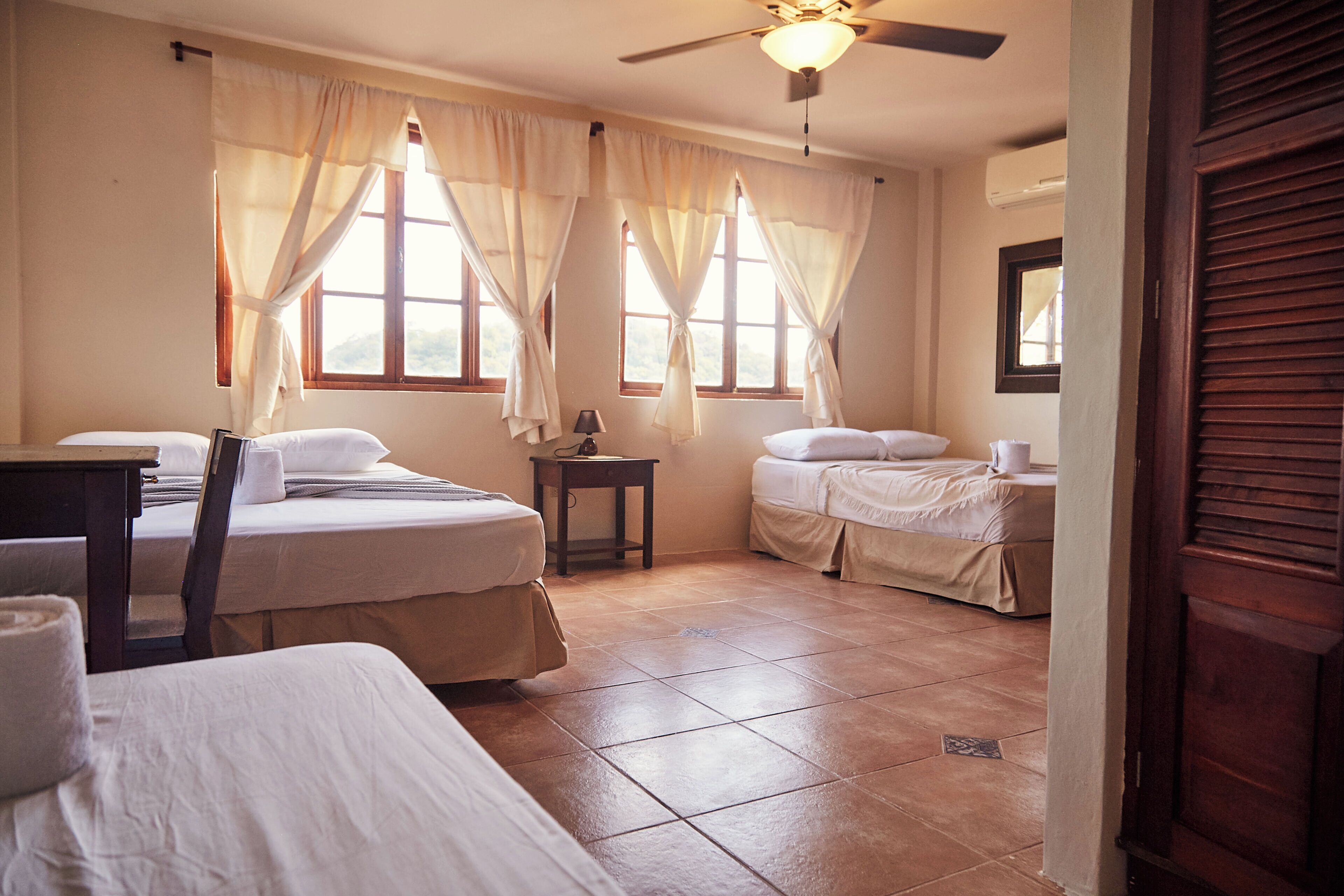 Ein Hotelzimmer mit zwei Betten, einem Deckenventilator und großen Fenstern mit Vorhängen.