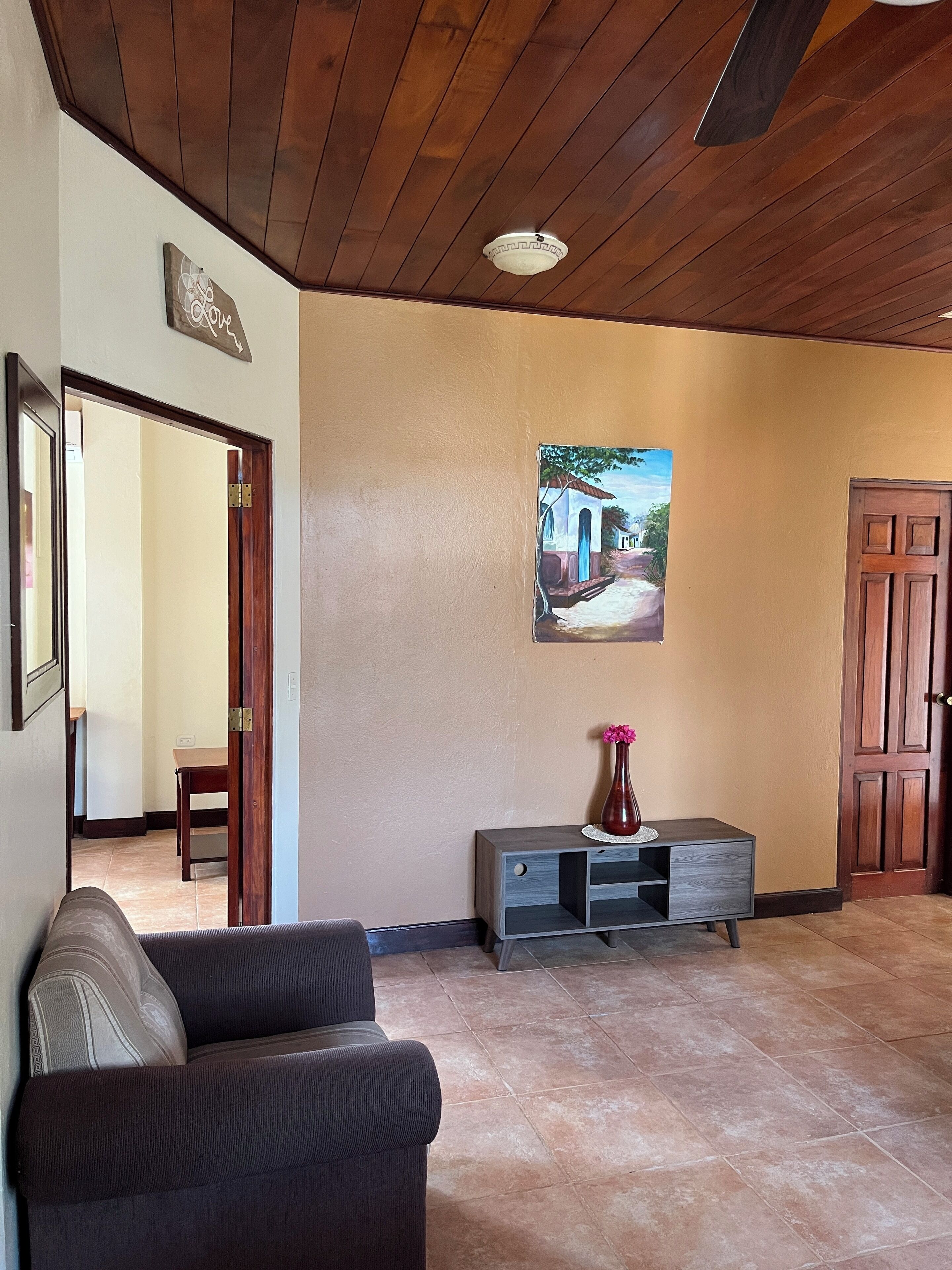 A spacious living area with a wooden ceiling, a painting of a street scene, a dark-colored sofa, and a wooden door.