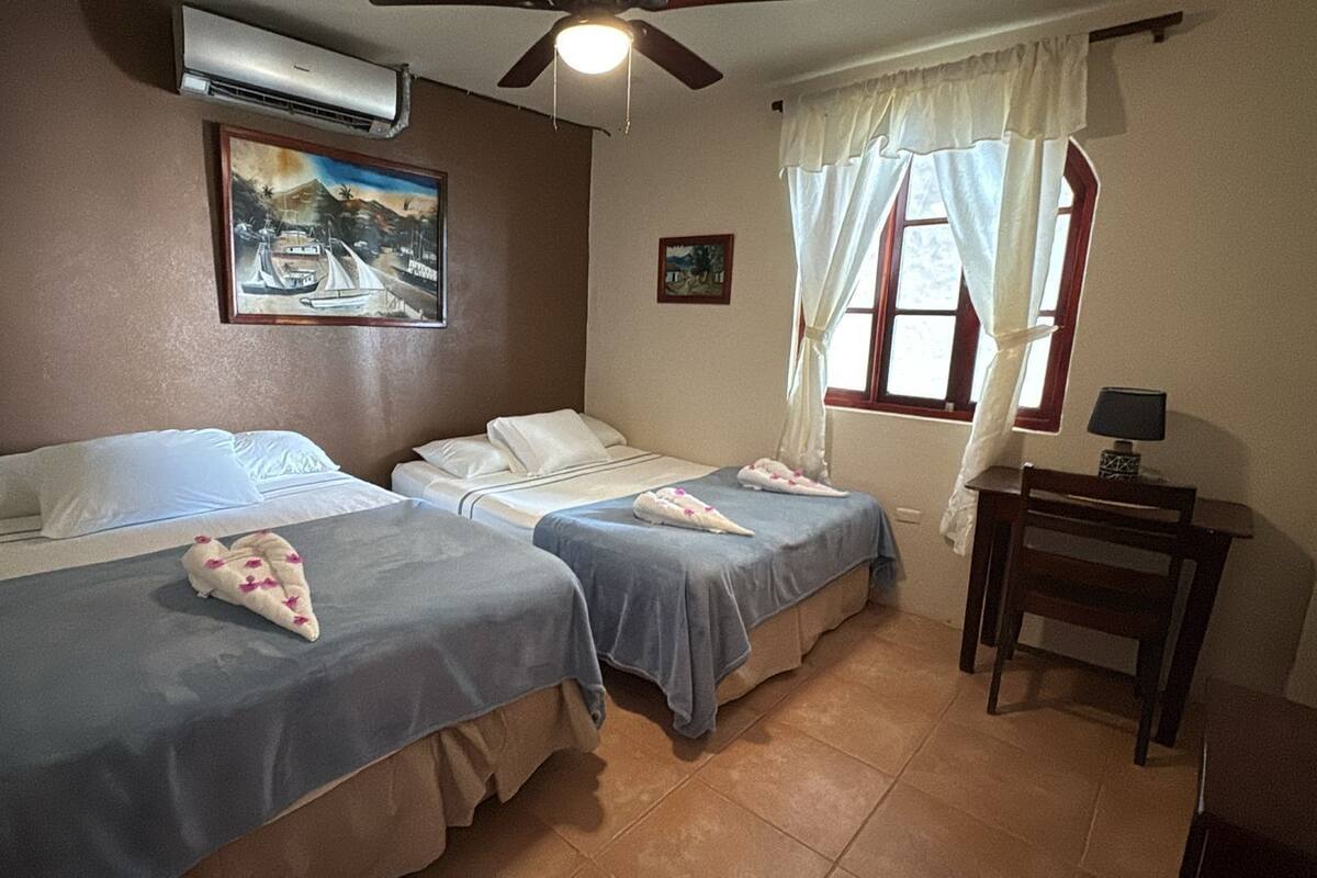 Une chambre d’hôtel avec deux lits, un ventilateur de plafond, un climatiseur, une fenêtre avec des rideaux et une petite table avec une lampe.