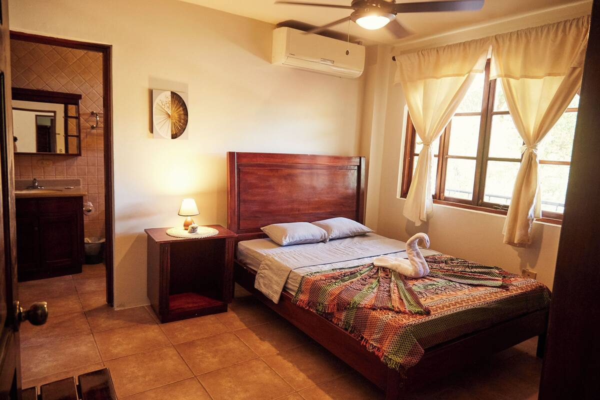 Une chambre avec un lit en bois, une table de chevet avec une lampe, un climatiseur fixé au mur et une fenêtre avec des rideaux.