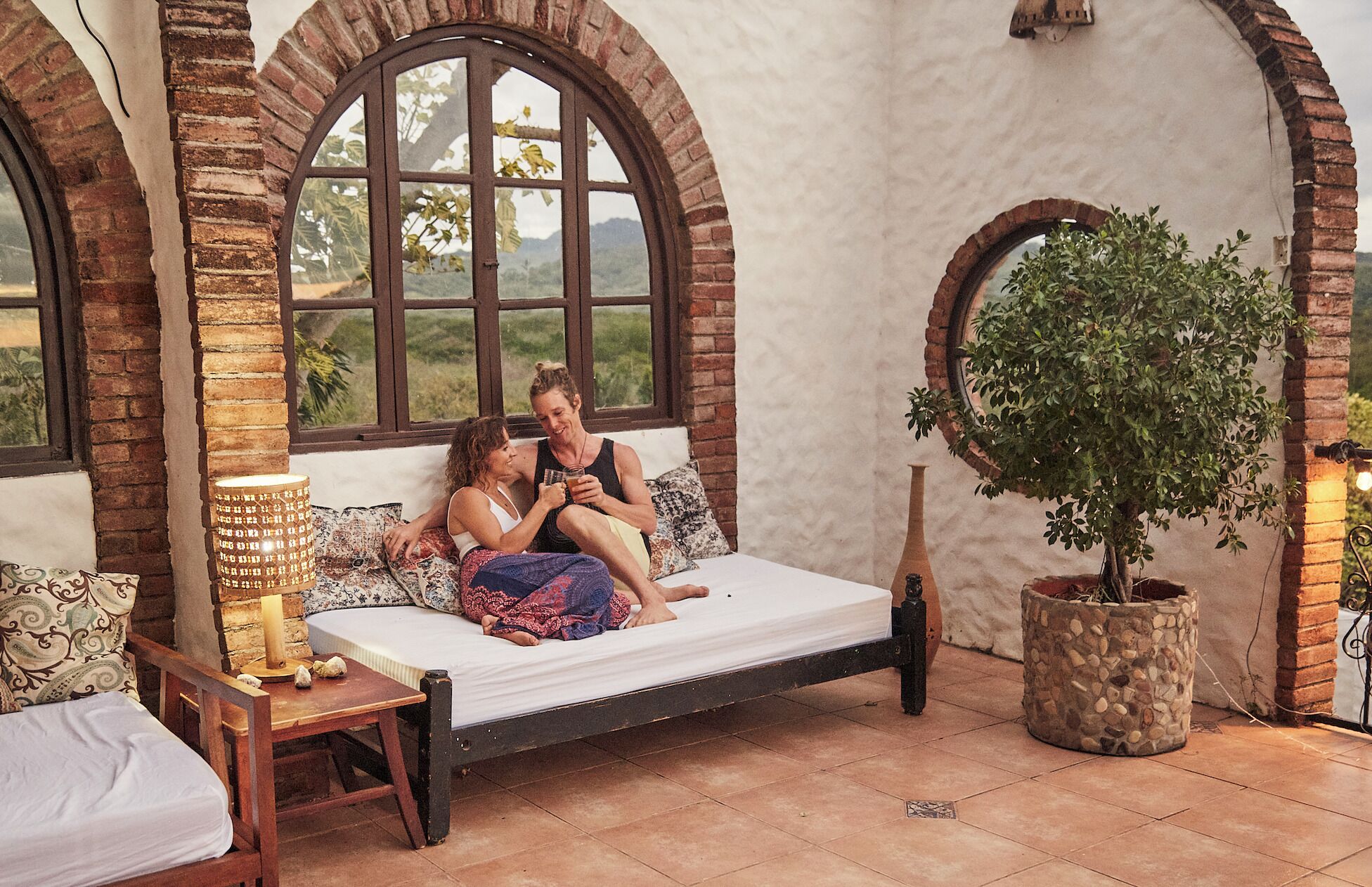 A rustic room with a wooden bed, a stone planter, and arched windows overlooking a landscape.