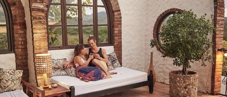 A rustic room with a wooden bed, a stone planter, and arched windows overlooking a landscape.
