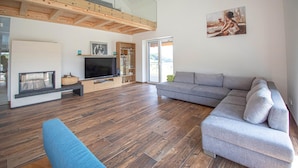 A modern living room with a large grey sectional sofa, a blue armchair, a flat-screen TV, and a wooden ceiling. - Ferienhaus in Kapfenstein (Kapfenstein)