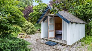 A small, single-room cabin with a wooden door and a window, surrounded by lush greenery and a gravel path. - 2 bedroom pet friendly home in Glesborg (Glesborg)