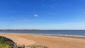 Une plage avec un ciel dégagé, une mer calme et une côte lointaine parsemée de bâtiments.