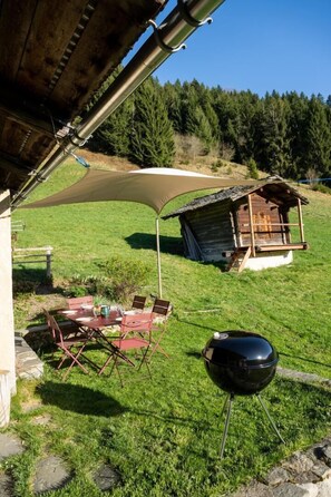 Ett takförsett utomhusområde med ett bord och stolar, en grill och en liten trähusliknande konstruktion på en gräsyta.