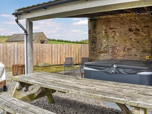 A wooden picnic table with a hot tub under a pergola, surrounded by a stone wall and a wooden fence.