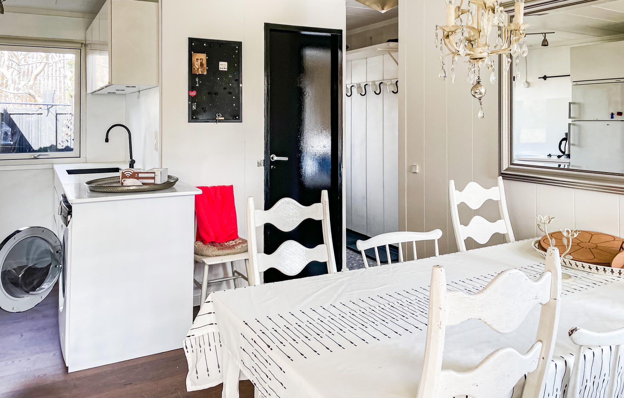 Una cocina con lavadora, una mesa de comedor con sillas blancas, una araña y una puerta negra con un tablón de anuncios.