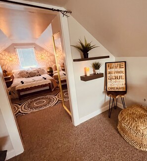 A cozy bedroom with a bed, a bedside table, a lamp, a window with blinds, a wall-mounted shelf with a plant, and a motivational poster.