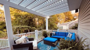 A covered deck with outdoor furniture, including a sofa and chairs, surrounded by trees and a white fence.