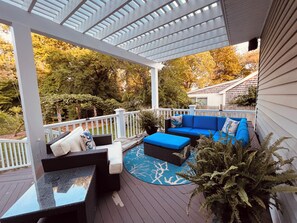 A covered deck with outdoor furniture, including a sofa and chairs, surrounded by trees and a white fence.