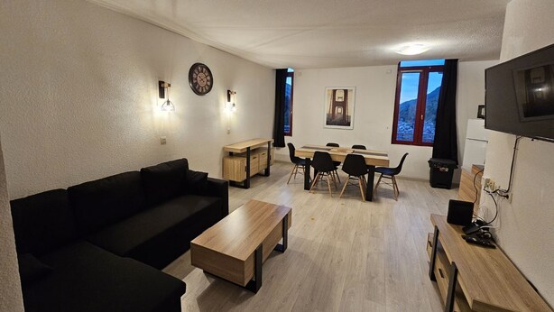 A modern living room with a sofa, coffee table, dining table, and chairs. There is a TV, a clock, and artwork on the walls.