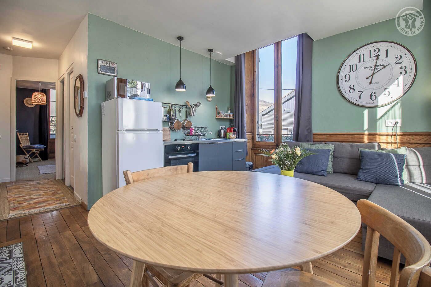 A modern kitchen with a refrigerator, oven, and hanging pots. There is a dining area with a round wooden table and chairs. A large clock is on the wall, and a sofa is in the background.