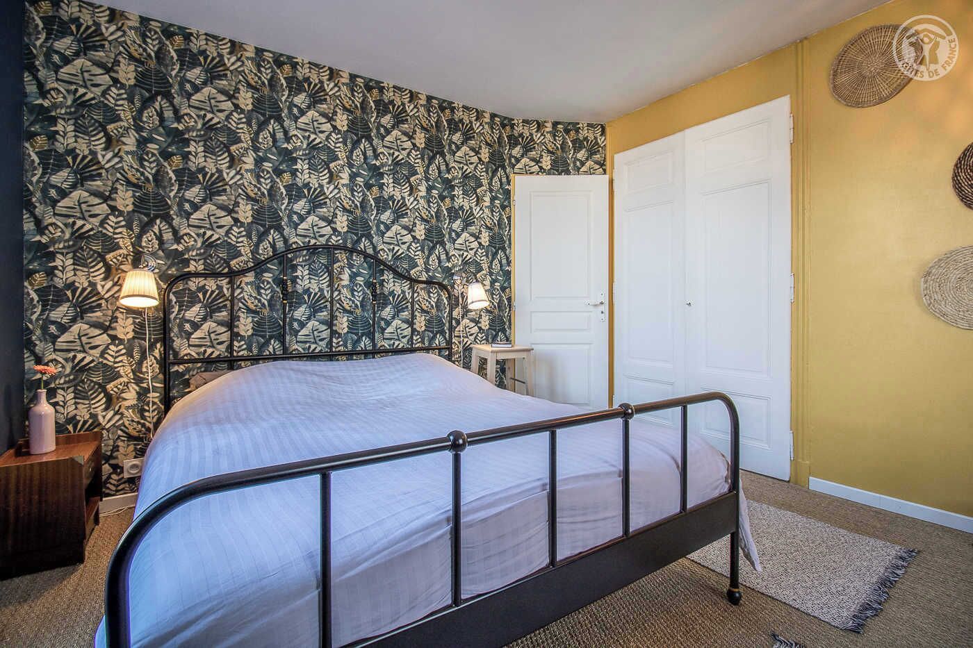A bedroom with a bed, a nightstand, a lamp, a door, and a wall with a leaf pattern.