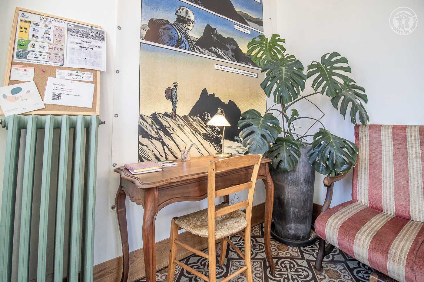 A cozy room with a wooden desk, a chair, a radiator, a large potted plant, and a striped sofa.