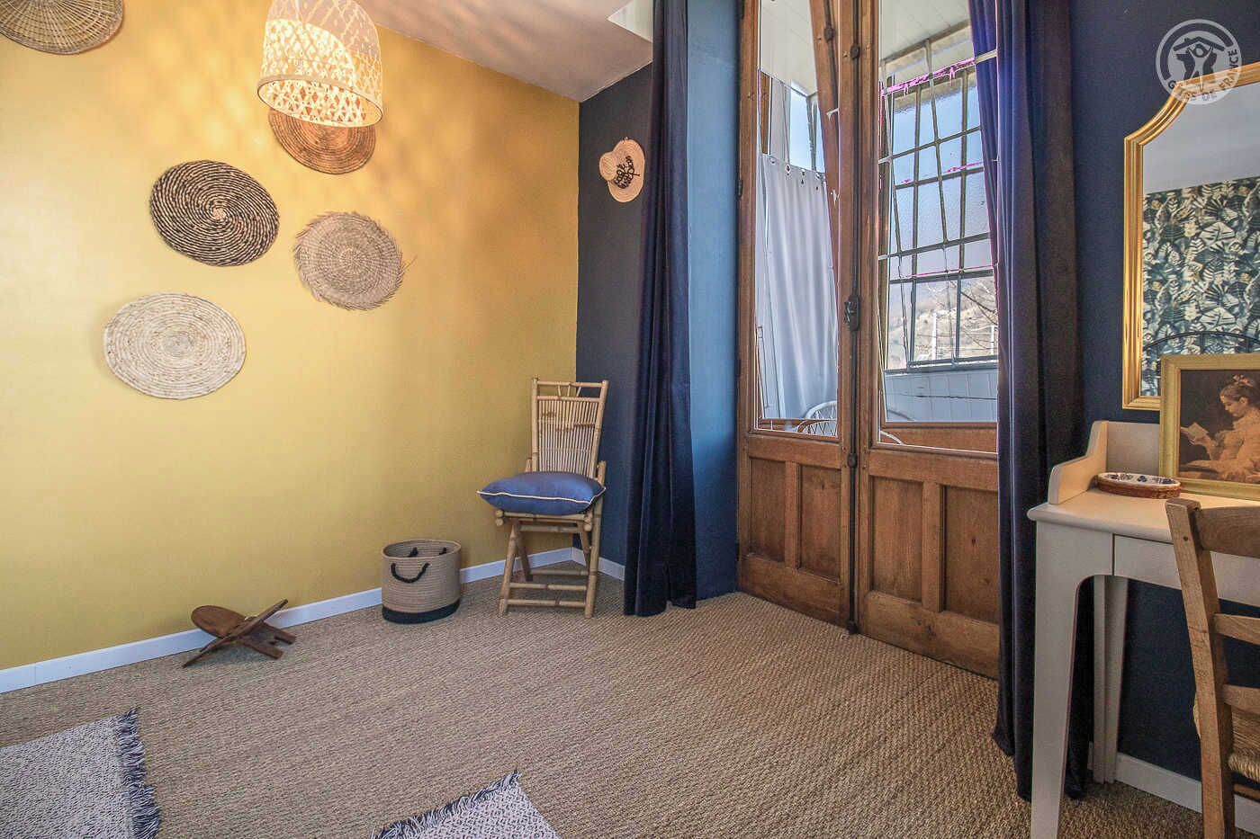 A room with a yellow wall, a wooden chair, a small table with a picture frame, and a window with curtains.