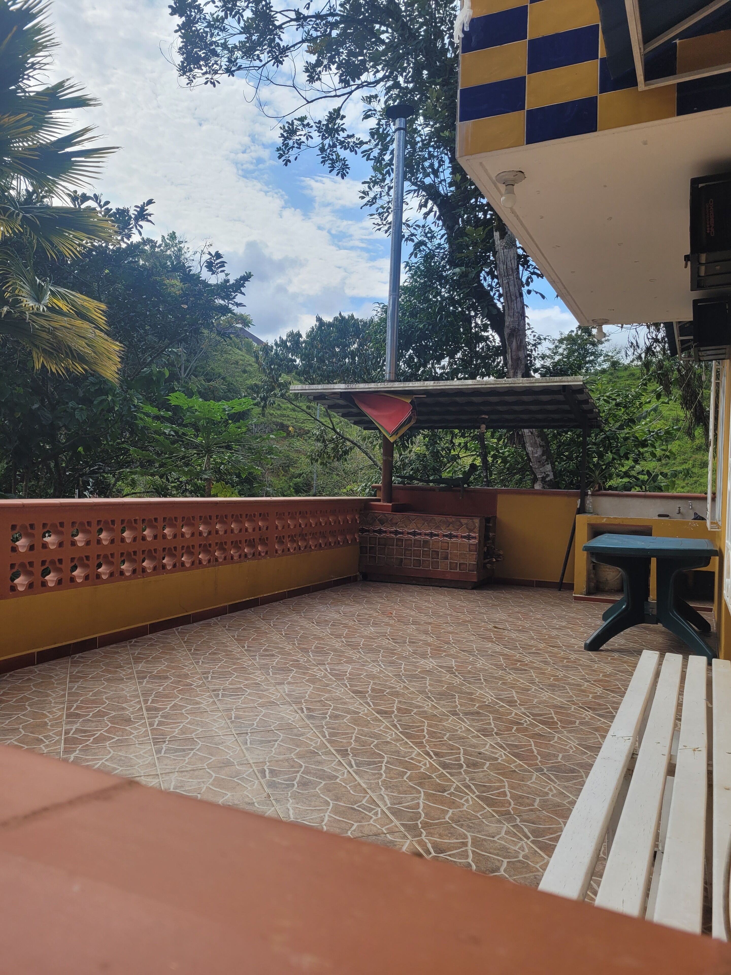 Un espacio al aire libre techado, con suelo de baldosas, una cerca roja y amarilla, y una mesa con un banco.