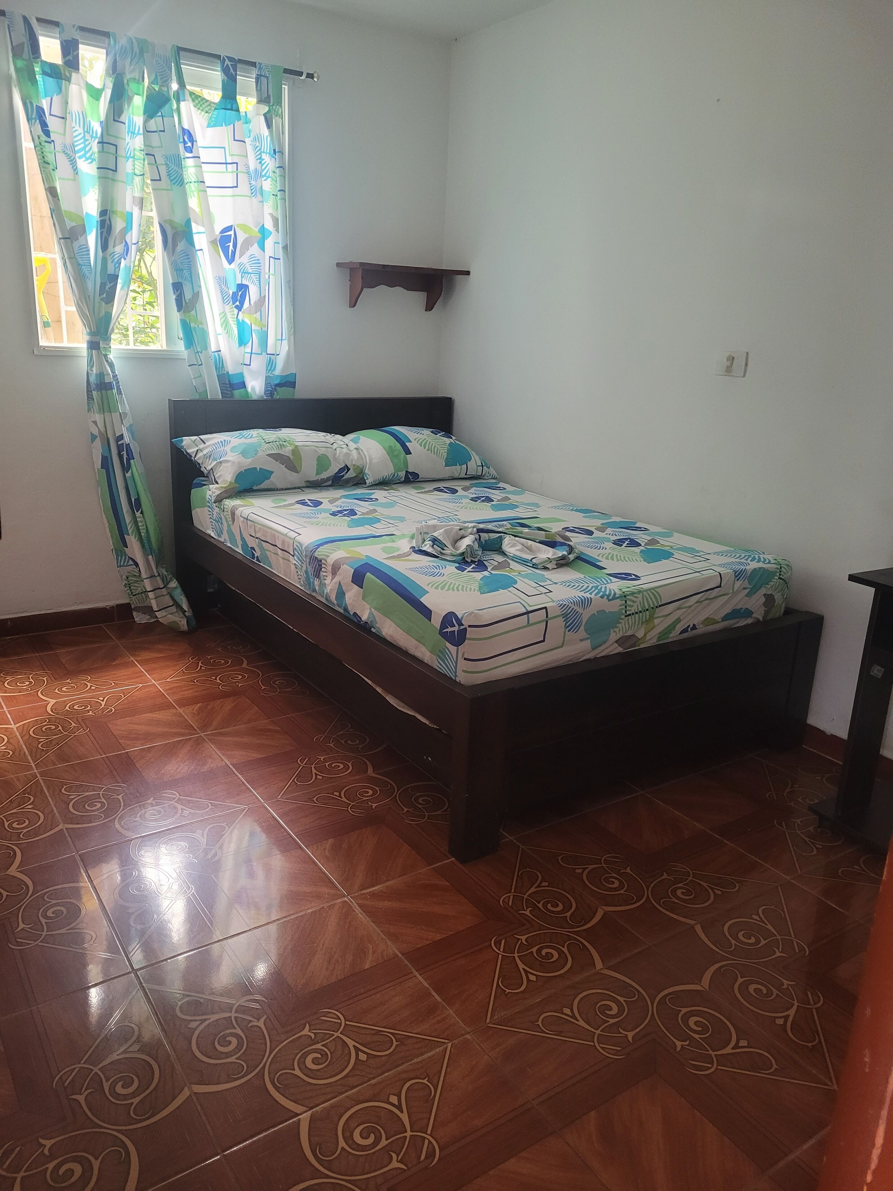 Quarto com cama, cortinas estampadas, prateleira pequena e chão de azulejos.