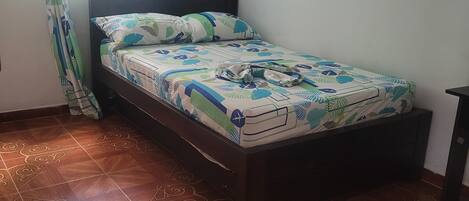 Quarto com cama, cortinas estampadas, prateleira pequena e chão de azulejos.