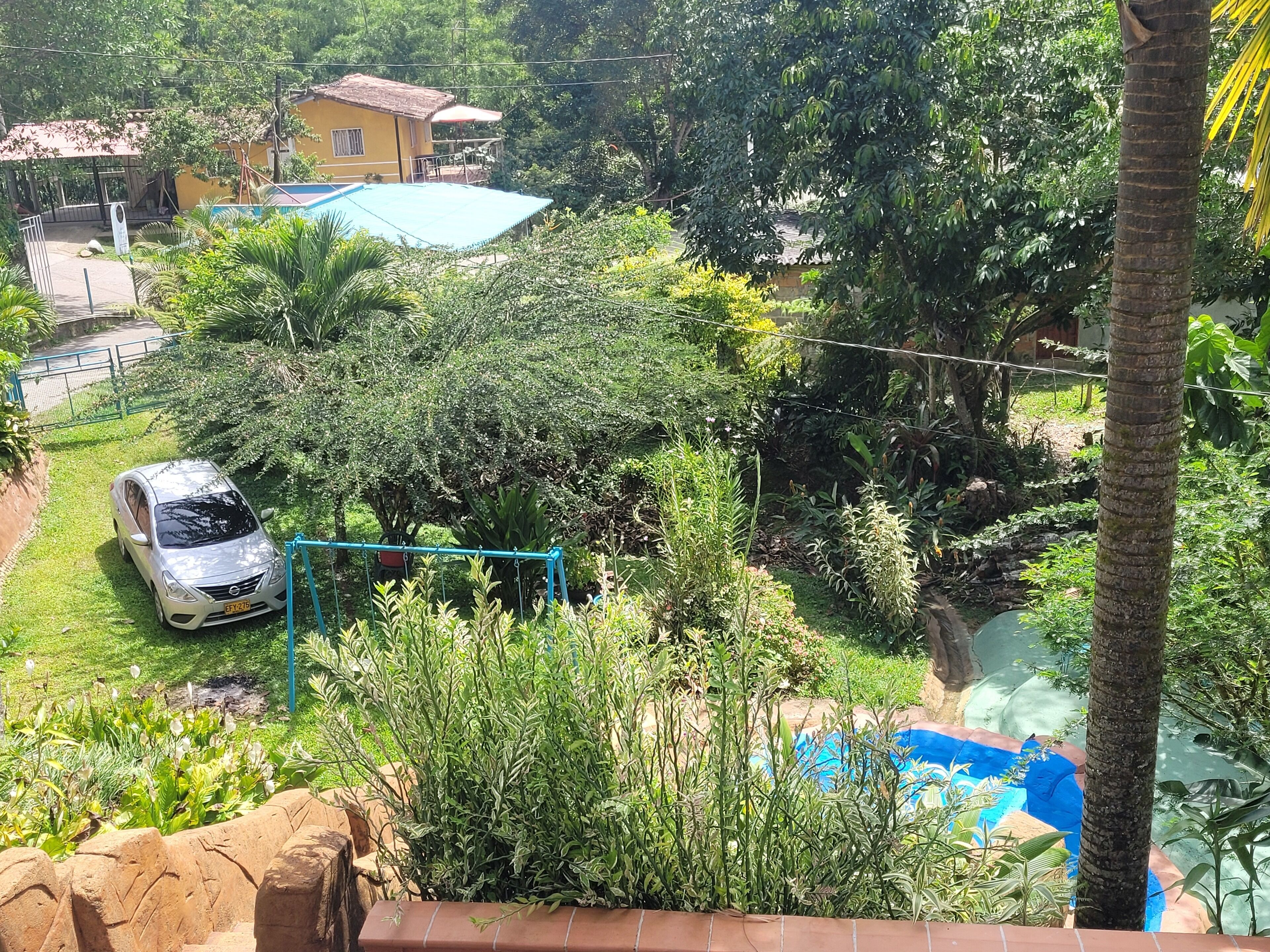 Ett bostadsområde med en parkerad bil nära en blå gunga, omgivet av frodig grönska och en liten pool.
