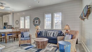A spacious living room with a blue sofa, wicker chairs, and a round coffee table. There is a kitchen in the background with white cabinets and a microwave. - Breez In TC 10304 (Port Aransas)