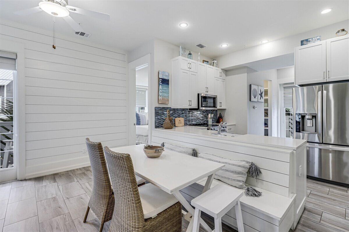 Una cocina moderna con muebles blancos, un refrigerador de acero inoxidable y un comedor con sillas de mimbre y una mesa blanca.
