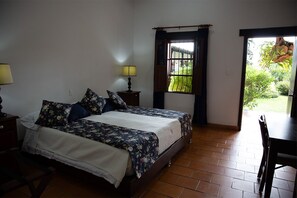 A bedroom with two beds, a nightstand with a lamp, a window with a view of greenery, and a door leading to an outdoor area. - Small boutique Hotel  on Ginebra Valle del Cauca home of the Mono Nunez festival (Ginebra)