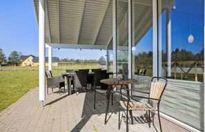Une terrasse couverte, aménagée avec du mobilier d’extérieur, une grande baie vitrée et une vue sur une maison jaune en arrière-plan.