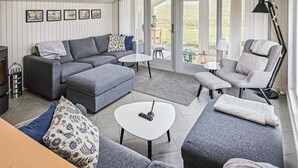 Un salón acogedor con un sofá gris, una mesa de centro blanca y un sillón gris. En la pared hay cuadros enmarcados, una mesita con una lámpara, y vistas al exterior a través de amplias puertas de cristal.