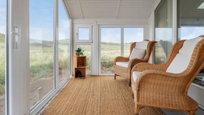 Una habitación con dos sillones de mimbre, un pequeño estante de madera con una planta y una copa de vino, y vistas a un paisaje con césped a través de amplias ventanas.