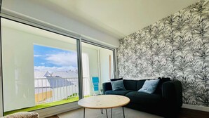 Ein modernes Wohnzimmer mit einer großen Schiebeglastür, die auf einen Balkon führt, einem dunkelgrünen Sofa, einem kleinen runden Tisch und einer gemusterten Tapete.