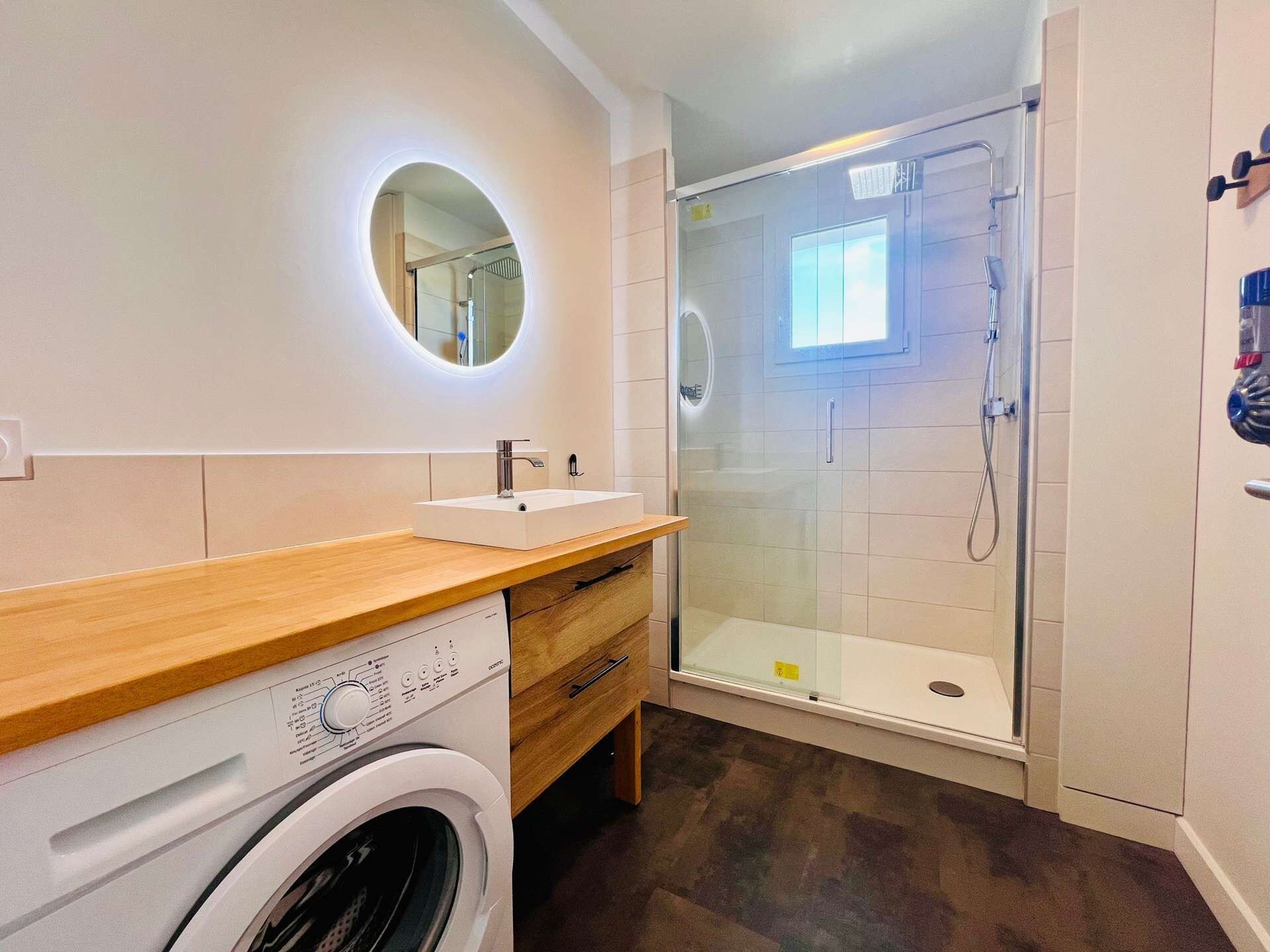 A modern bathroom with a built-in washing machine, a glass-enclosed shower, and a round mirror with integrated lighting.