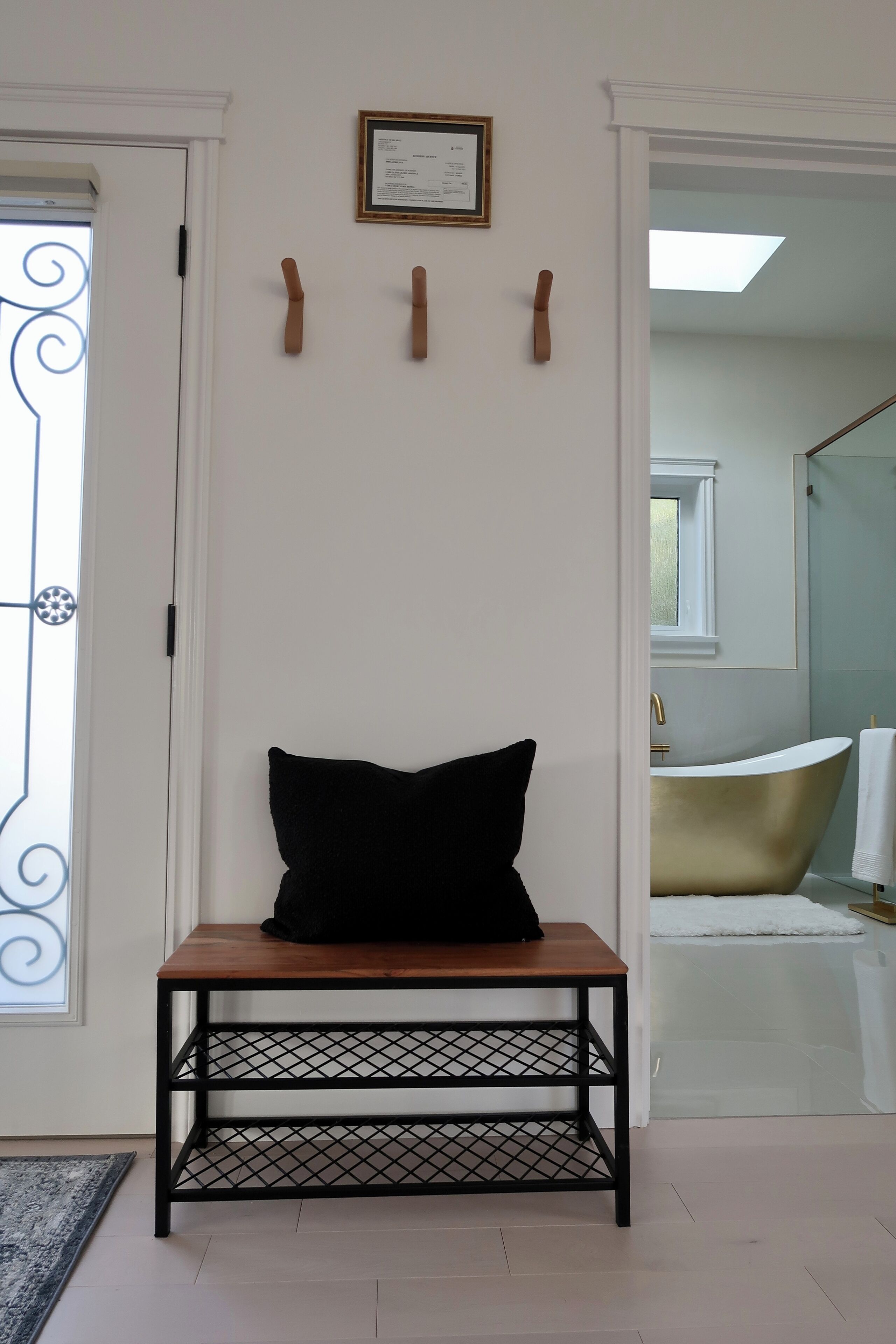 Ein Zimmer mit einer hölzernen Bank, einem schwarzen Kissen, einem gerahmten Zertifikat an der Wand und einem Blick ins Bad mit einer Badewanne und einem Handtuchhalter.