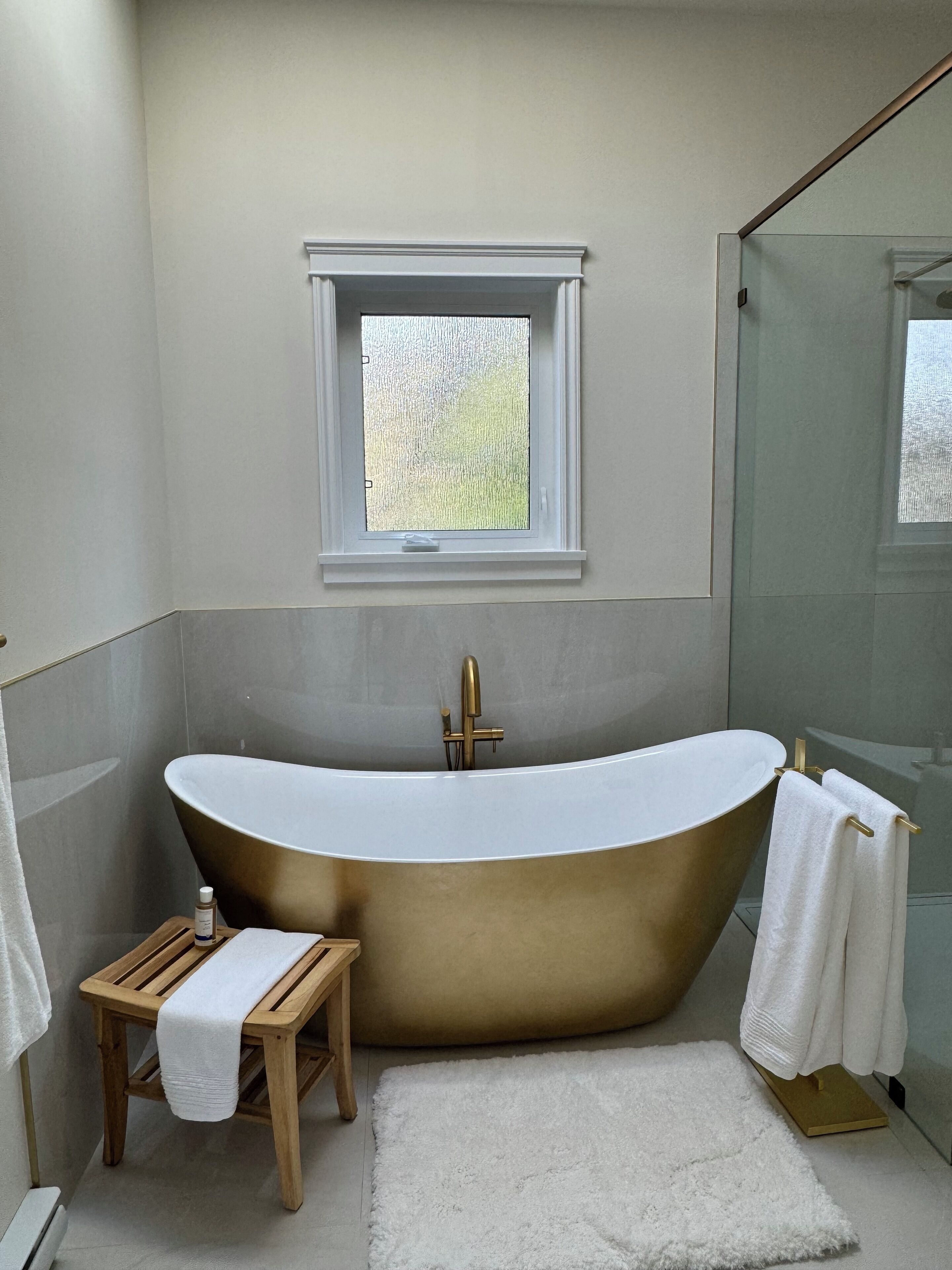 A modern bathroom with a freestanding bathtub, a small wooden stool, a white rug, and a window with a view of greenery.