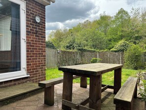 Een houten patio tafel en bankjes in een tuin, met een bakstenen muur en een houten omheining op de achtergrond.