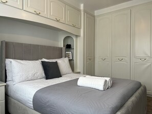 Een opgemaakt bed met een grijs hoofdeinde, wit beddengoed en een grijze plaid.
