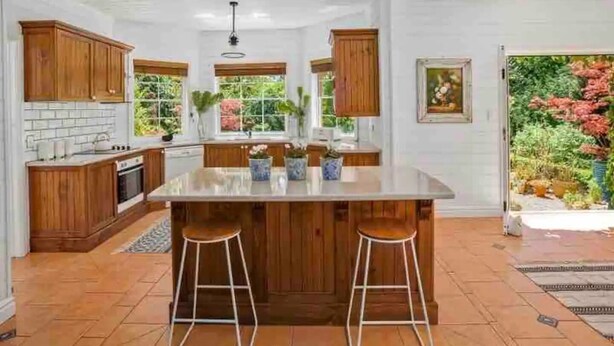 A modern kitchen with wooden cabinets, a large island with stools, and a view of a garden. - Stunning Highlands Manor-Accommodates 2-12 people  (Mittagong)