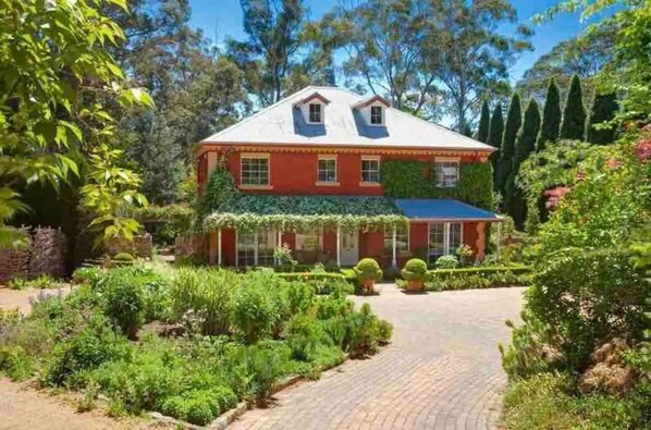 A red house with a white roof, surrounded by a garden with various plants and a paved pathway leading to the entrance. - Stunning Highlands Manor-Accommodates 2-12 people  (Mittagong)