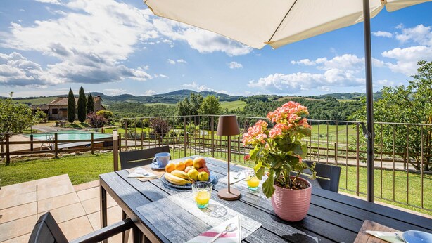 Een terras met een ontbijttafel, een zwembad en een mooi uitzicht op heuvels en een huis.