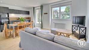 Een moderne woonkamer met een grijze bank, eettafel en stoelen. Er is een keuken met een magnetron, oven en kastjes. Een televisie is bevestigd aan een zwarte standaard, en een raam biedt uitzicht op bomen.