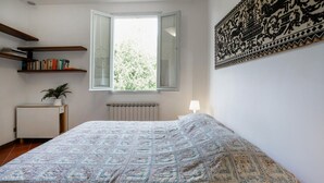 A bedroom with a bed, a radiator, a window with an open shutter, a bedside table with a lamp, a potted plant, and a wall with a decorative tapestry. - Nice apartment in Firenze with WiFi (Firenze)