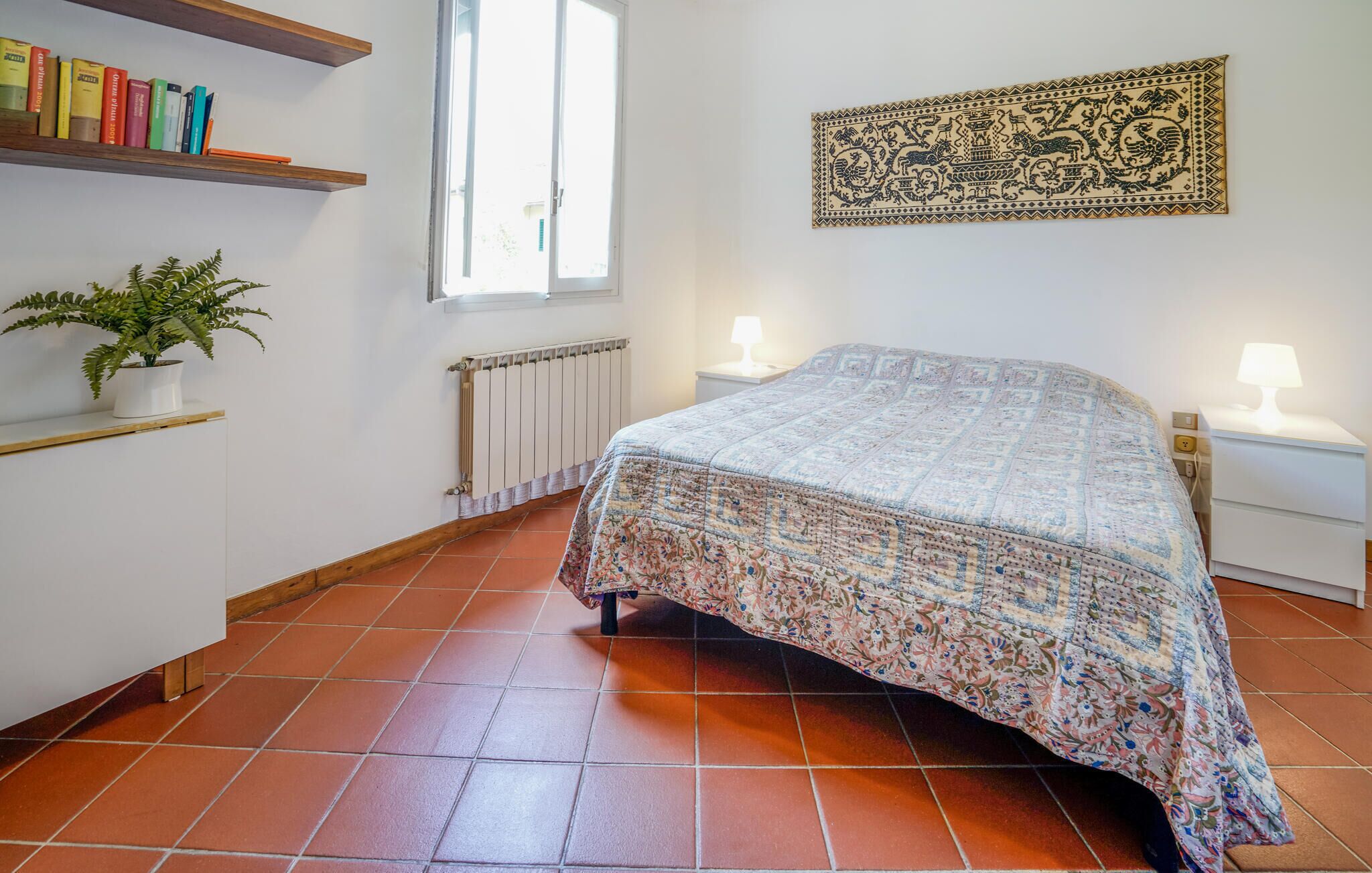 A bedroom with a bed, a radiator, a window, a shelf with books, a plant, and a tapestry on the wall.