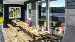 Un área de comedor al aire libre con una mesa de madera dispuesta para dos personas, rodeada de amplios ventanales que ofrecen vistas a un cuerpo de agua y vegetación.