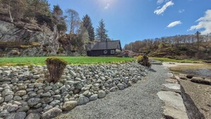 Una casa con un camino de piedra que lleva a una zona rocosa, rodeada de árboles y un cielo despejado.