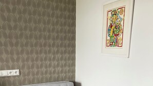 Harmaa sohva, jossa on koristetyyny, kuviollinen tapetti, kehystetty taideteos ja pistorasia seinässä.
